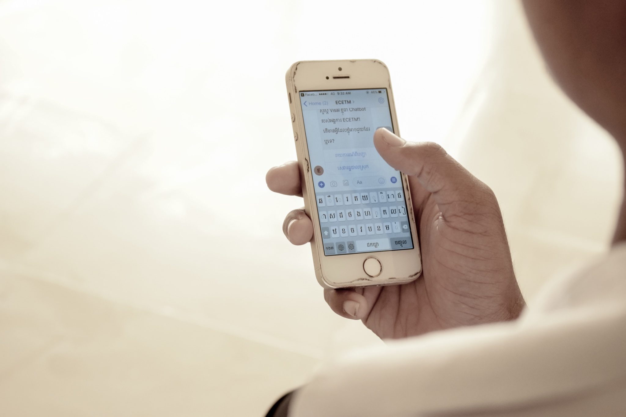 A hand holds an iPhone showing the chatbot conversation in Khmer, with a Khmer keyboard open in Messenger